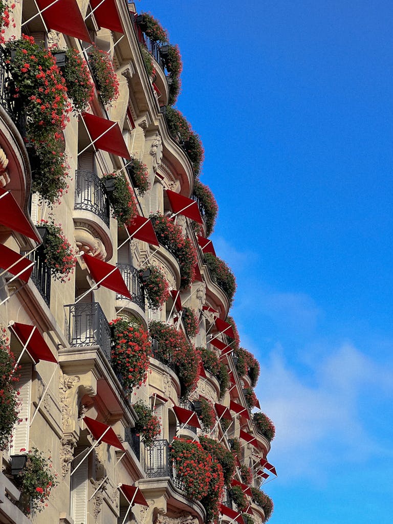 Luxury Parisian hotel exterior with red awnings and floral balconies against clear blue sky.